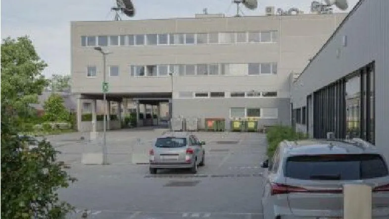 Gebäude mit moderner Architektur und Satellitenschüsseln auf dem Dach, umgeben von einem Parkplatz und Grünflächen.