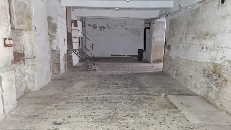 Großer, leerer Raum im Kellergeschoss mit sichtbaren Betonbalken und einer Metalltreppe, in sanierungsbedürftigem Zustand.