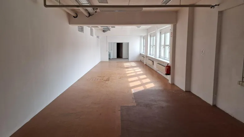 Geräumiger, leerer Büroraum mit vielen Fenstern, die viel Tageslicht hereinlassen und freiliegenden Deckeninstallationen.