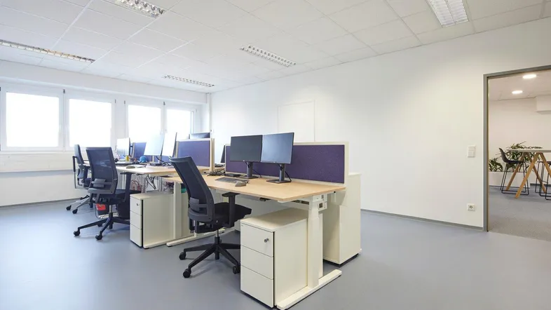 Helles und modernes Büro mit mehreren Arbeitsplätzen, großen Fenstern und grauem Bodenbelag.