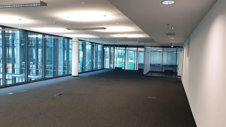 Geräumiger, leerer Büroraum mit großen Glasfronten und dunklem Teppichboden, ideal für moderne Arbeitsumgebungen.