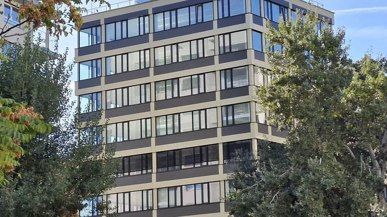 Modernes Bürogebäude mit vielen Fenstern und umgeben von grünen Bäumen unter blauem Himmel.