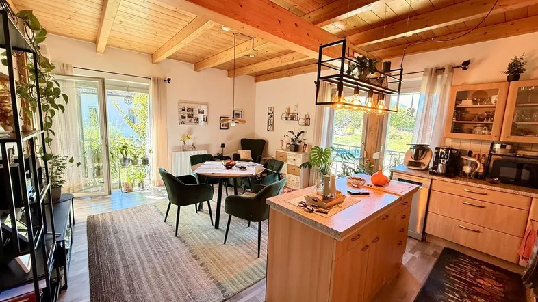 Heller offener Wohn-Essbereich mit Holzbalkendecke, integrierter Küche und Zugang zum Balkon.