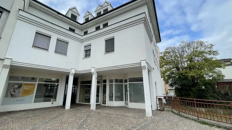 Modernes Geschäftsgebäude mit weißer Fassade, großen Fenstern und gepflastertem Vorplatz unter bewölktem Himmel.
