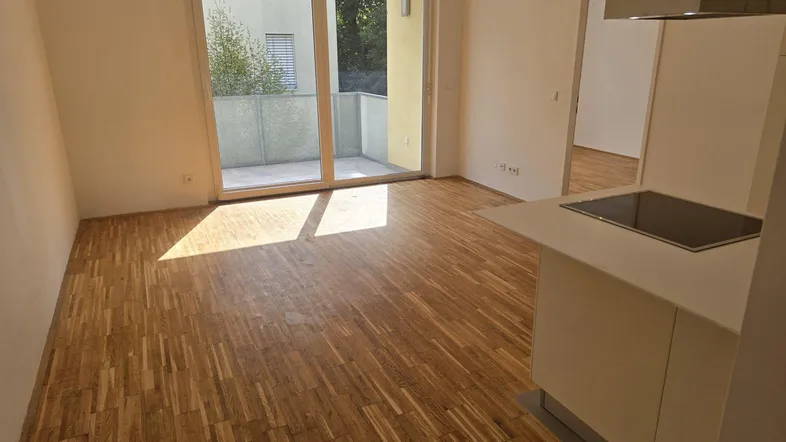 Heller Wohn-Essbereich mit Parkettboden, Zugang zum Balkon und integrierter Küchenzeile.