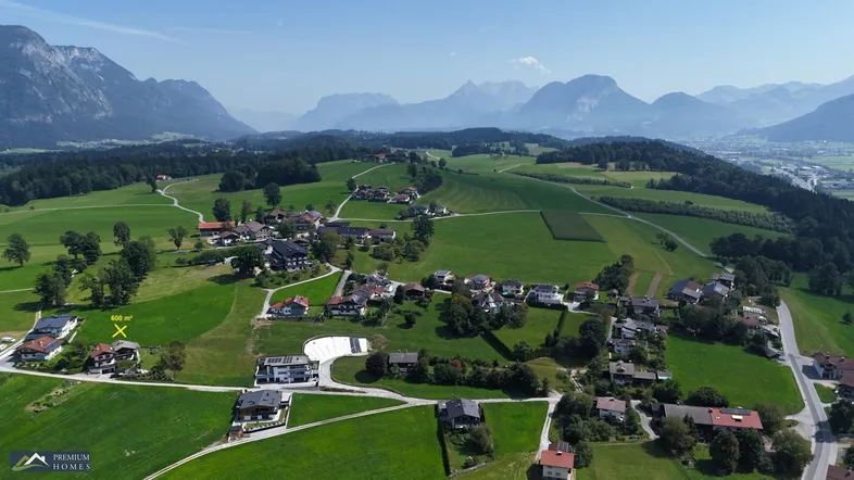 Luftaufnahme einer ländlichen Siedlung mit Häusern, grünen Feldern und majestätischen Bergen im Hintergrund.