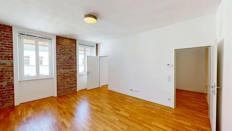 Heller Wohnraum mit freigelegten Ziegelwänden, Parkettboden und großen Fenstern.