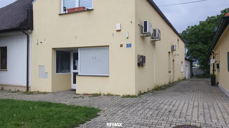 Gelbes Gebäude mit Schaufenster und Eingangsbereich, gepflasterter Vorplatz und Klimaanlagen an der Fassade.