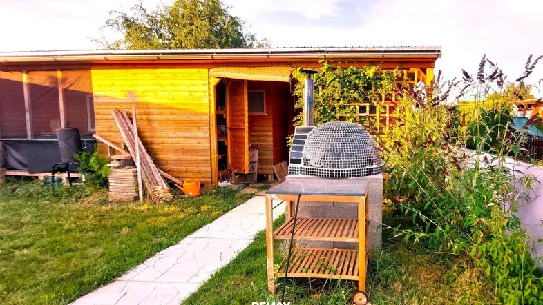 Hölzernes Gartenhaus mit einem gemauerten Pizzaofen und einem gepflasterten Weg im Grünen.