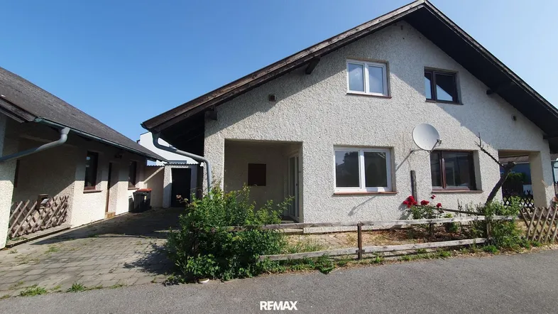 Einfamilienhaus mit heller Fassade, Satteldach und angrenzendem Nebengebäude unter blauem Himmel.