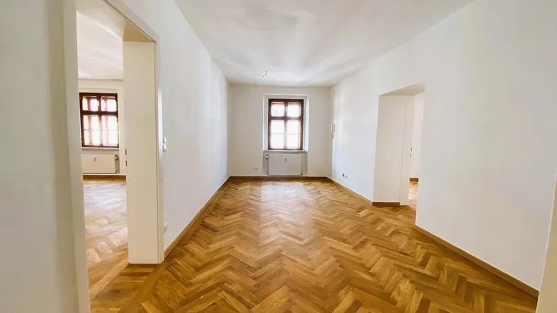 Helles, leeres Zimmer mit Fischgrätparkett und zwei Fenstern, bereit zur individuellen Gestaltung.