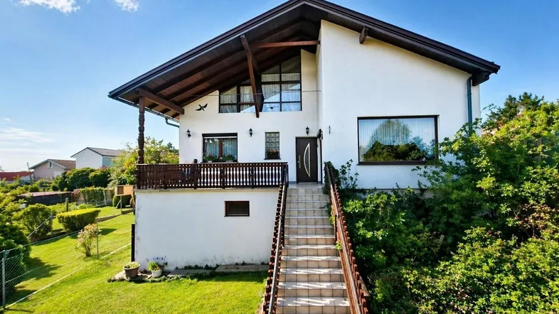 Weißes Einfamilienhaus mit Holzbalkon und langer Steintreppe zum Eingang.