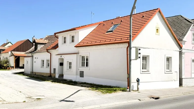 Weißes Wohnhaus mit rotem Ziegeldach und mehreren Fenstern an einer sonnigen Straße gelegen.
