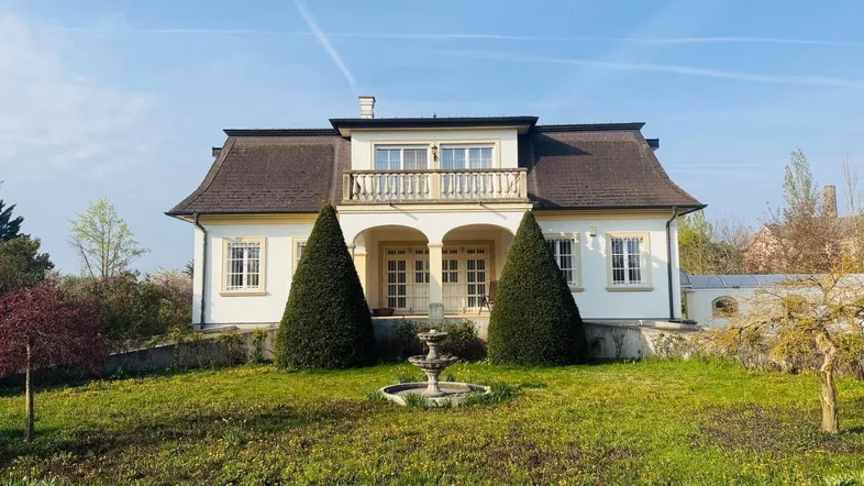 Elegante Villa mit gepflegtem Vorgarten und dekorativem Brunnen unter blauem Himmel.
