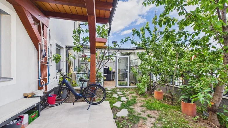 Gartenbereich mit gepflasterter Fläche, Fahrrad und einem verglasten Wintergarten oder Gewächshaus.