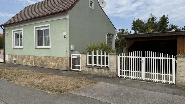 Ansicht des Hauses von der Straße mit Vorgarten, Zaun und einem einfachen Carport.