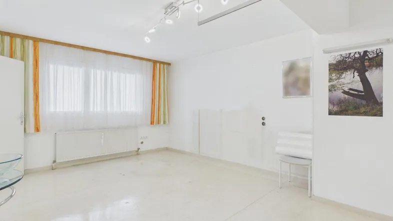 Helles, leeres Zimmer mit großen Fenstern, Heizkörper und schlichtem Bodenbelag.