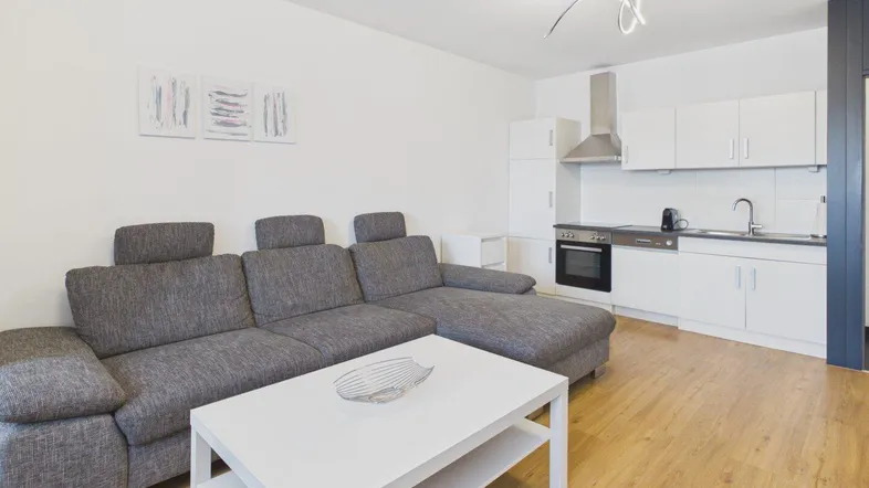 Heller offener Wohnbereich mit bequemer Couch, modernem Couchtisch und integrierter Küchenzeile mit Holzboden.