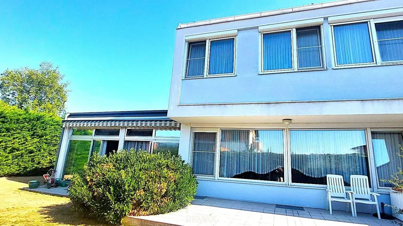 Modernes Haus mit großen Fenstern, einer Terrasse und einem gepflegten Gartenbereich mit Rasen und Hecke.