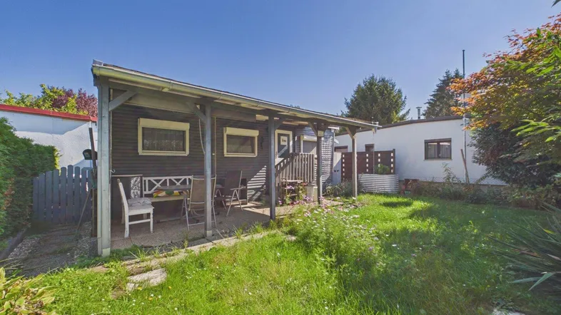 Kleines Haus mit überdachter Terrasse und Gartenbereich, umgeben von viel Grün.