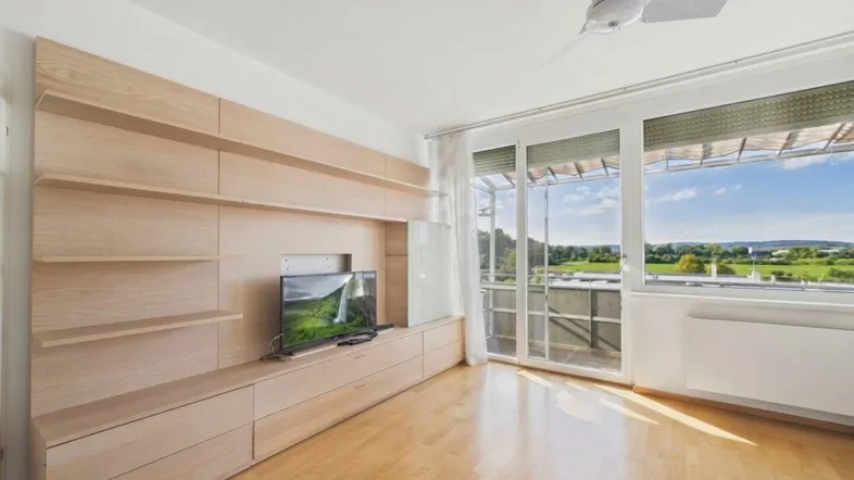 Helles Wohnzimmer mit integrierter Holzschrankwand, Zugang zum Balkon und weitem Blick ins Grüne.
