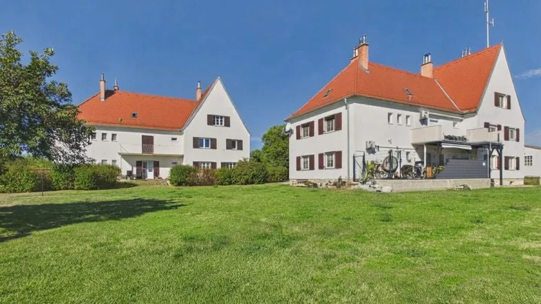 Zwei weiße Gebäude mit roten Dächern und einem weitläufigen grünen Rasen unter blauem Himmel.