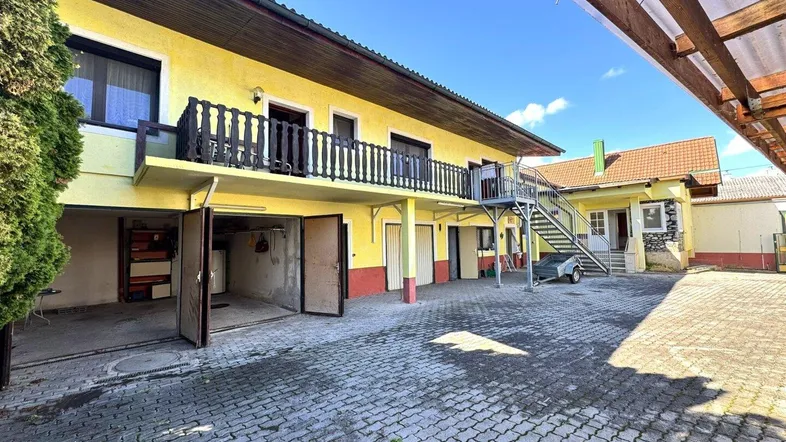 Gelbes Gebäude mit Garagen, externer Treppe und gepflastertem Vorplatz unter blauem Himmel.