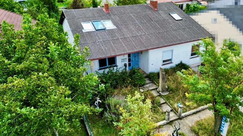 Haus mit Satteldach und mehreren Dachfenstern, umgeben von üppigem Grün und einem verwilderten Garten.