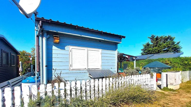 Kleines blaues Holzhaus mit Satellitenschüssel und weißem Zaun unter blauem Himmel.
