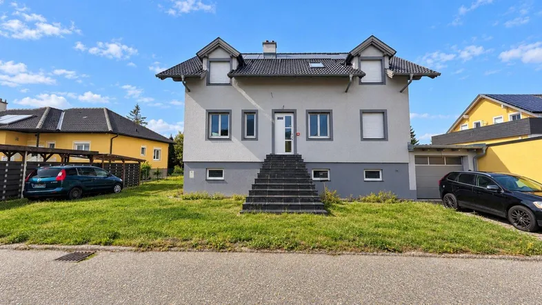 Graues Einfamilienhaus mit Satteldach und Vorgarten, von der Straße aus gesehen.
