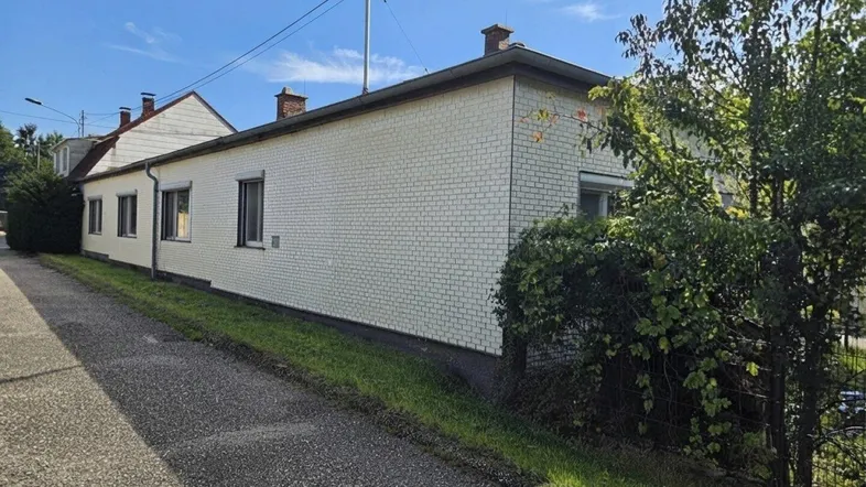 Seitenansicht eines Hauses mit weißer Ziegelfassade und grüner Bepflanzung unter blauem Himmel.