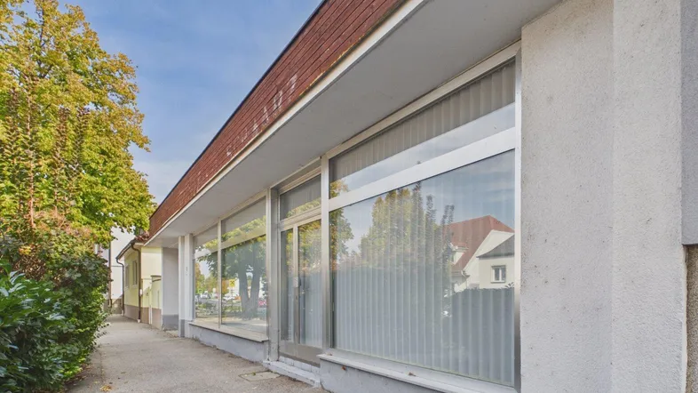 Ansicht der Geschäftsfassade mit großen Schaufenstern und einem Gehweg, umgeben von Bäumen und Sträuchern.
