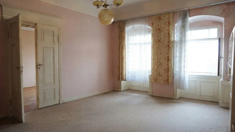 Geräumiges Zimmer mit rosa Tapete, Teppichboden und zwei hohen Bogenfenstern, bereit zur Renovierung.