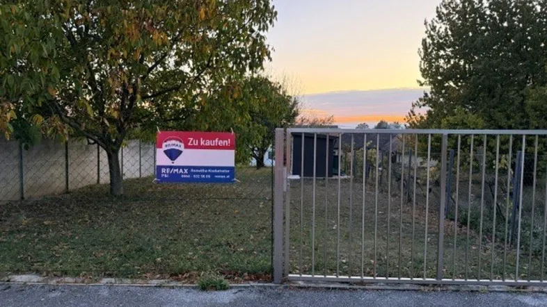 Grundstück mit Zaun und 'Zu kaufen!'-Schild bei Sonnenuntergang, umgeben von Bäumen und Natur.