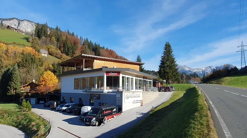 Modernes Geschäftsgebäude in alpiner Landschaft mit Parkplätzen und Bergblick unter blauem Himmel.