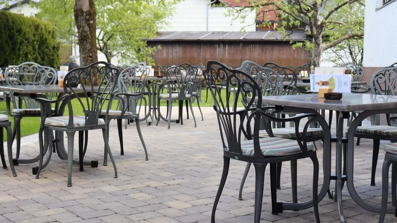 Eine gepflasterte Terrasse mit zahlreichen schmiedeeisernen Tischen und Stühlen, umgeben von Bäumen und Grünflächen.