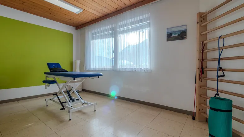 Heller Therapieraum mit Behandlungsliege, Sprossenwand und einer Akzentwand in frischem Grün.