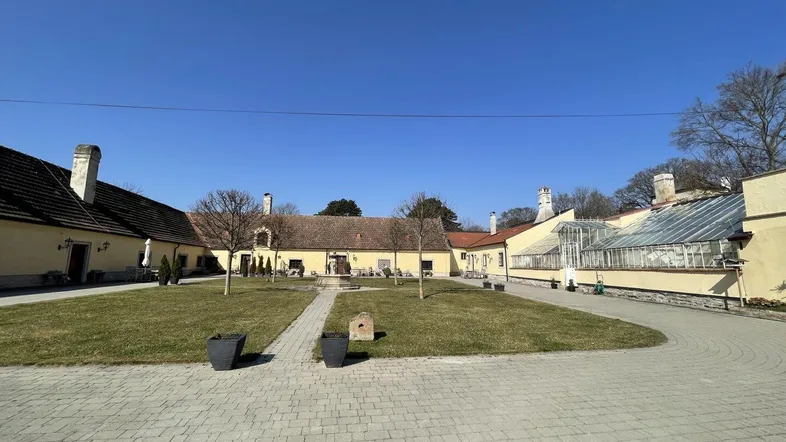 Weitläufige Außenansicht eines historischen Anwesens mit gepflastertem Hof, Rasenfläche und Gewächshaus unter blauem Himmel.