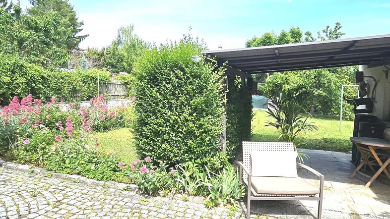 Gepflasterter Außenbereich mit Garten, Hecke und überdachtem Sitzplatz im Grünen.