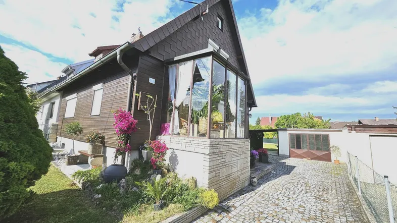 Hausfassade mit Wintergarten, gepflasterter Auffahrt und gepflegtem Vorgarten unter blauem Himmel.