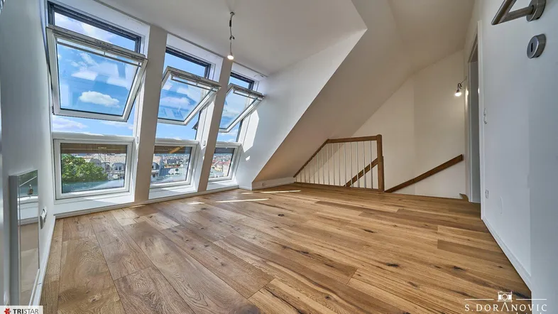 Heller Dachgeschossraum mit Holzboden, vielen Dachfenstern und Blick auf die Stadt.
