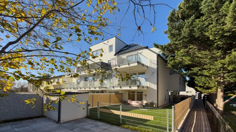 Modernes Mehrfamilienhaus mit Balkonen, kleiner Rasenfläche und gepflastertem Zugangsbereich unter blauem Himmel.