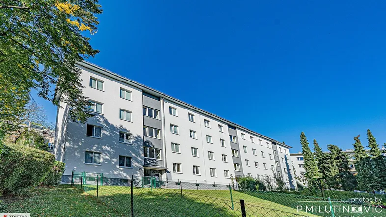 Mehrfamilienhaus mit weißer Fassade und grüner Umgebung unter blauem Himmel.