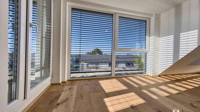 Helles Zimmer mit Holzboden und großen Fenstern, die viel Sonnenlicht hereinlassen.