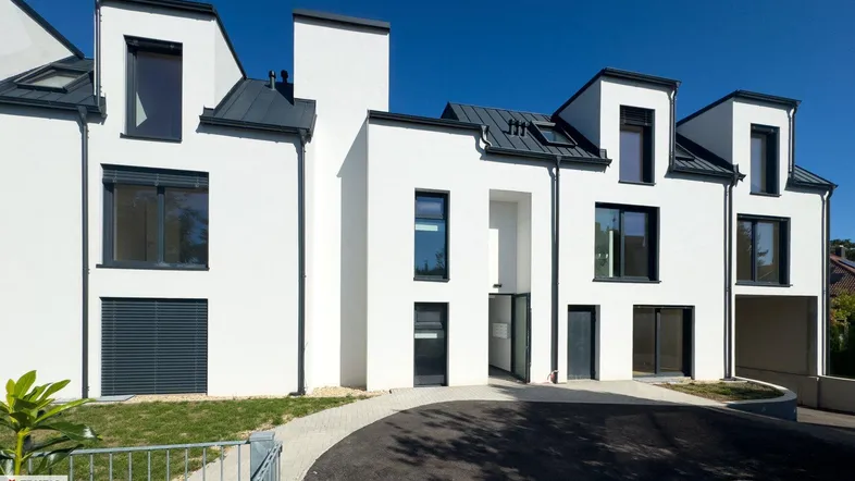 Moderne weiße Fassade eines Neubaus mit dunklen Fenstern und Rollläden, umgeben von Grünflächen.