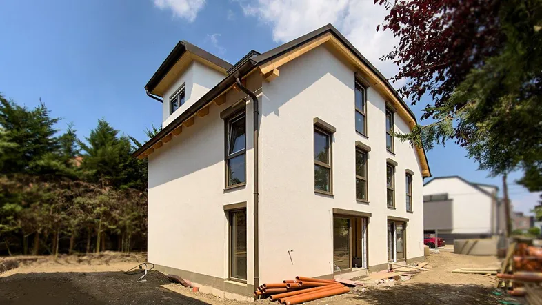 Modernes Einfamilienhaus im Bau mit weißer Fassade und dunklen Fenstern unter blauem Himmel.