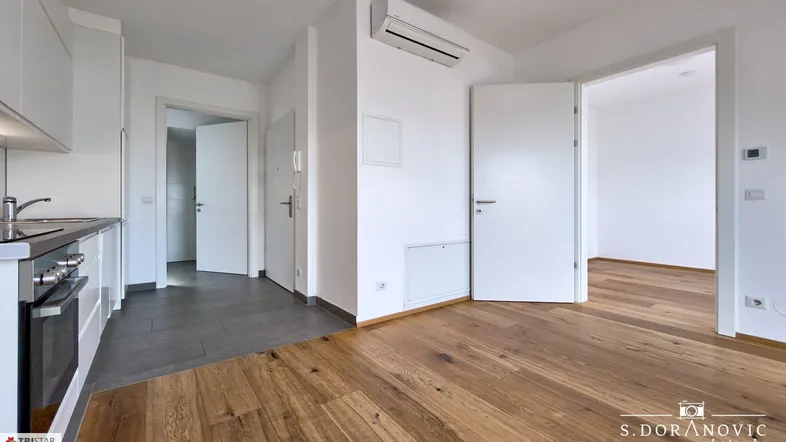 Heller Küchenbereich mit Holzboden, Klimaanlage und Zugang zu weiteren Räumen.
