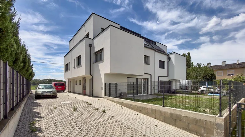 Modernes weißes Gebäude mit klaren Linien, gepflasterter Auffahrt und kleinem Gartenbereich unter blauem Himmel.