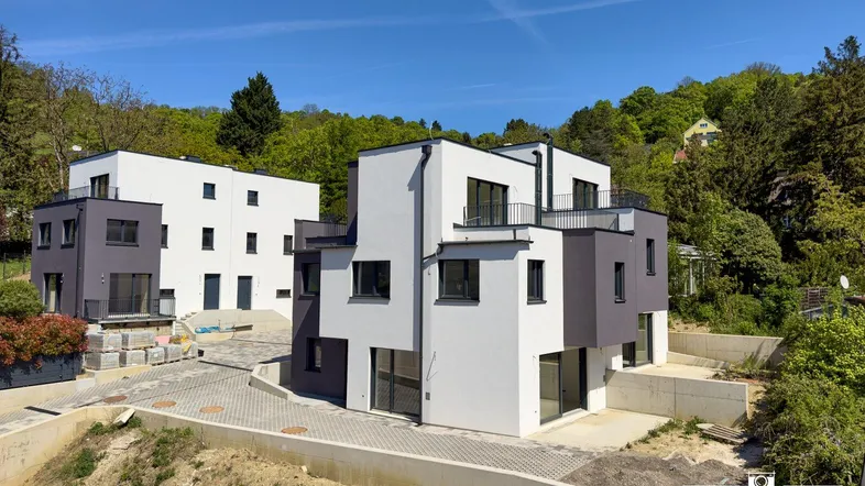 Moderne Neubauten mit weißer und grauer Fassade, eingebettet in eine grüne Hügellandschaft.