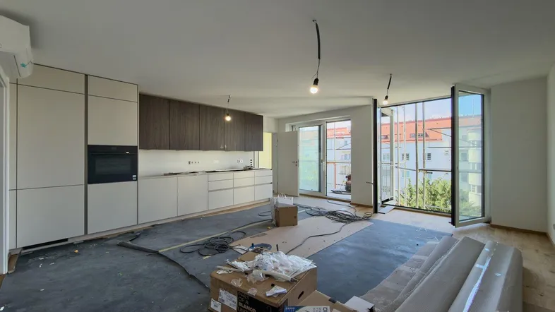 Offener Wohnbereich mit integrierter Küche, großen Fenstern und Zugang zum Balkon, noch im Bau.